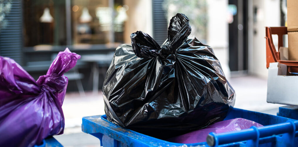 Black garbage bag atop a blue bin in an urban alleyway, awaiting disposal service.