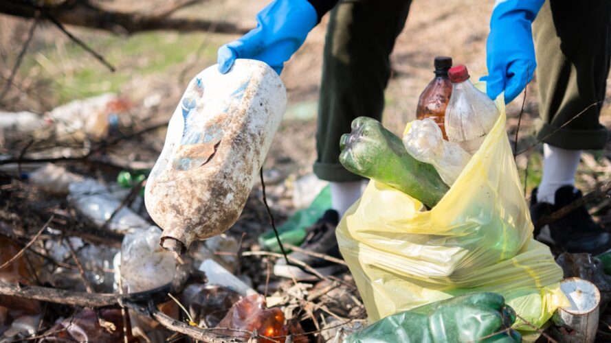 Wearing blue gloves, a person collects plastic bottles and trash into a yellow bag outdoors, promoting responsible trash disposal.